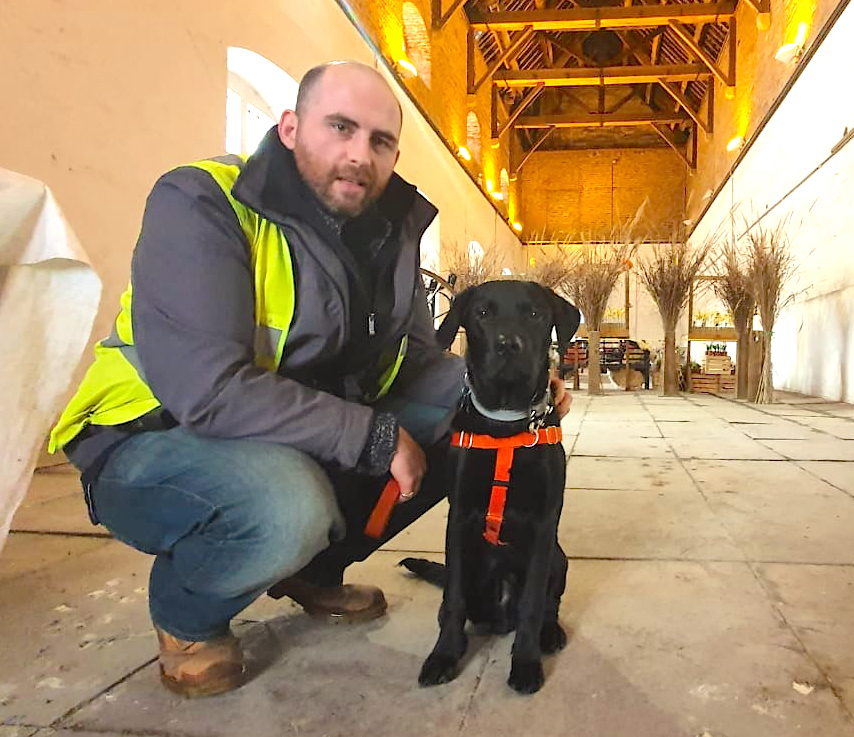 Archaeologist James Miles squats next to a sitting black labrador puppy wearing a bright orange harness inside an old building with a high ceilling, wooden rafters, and stone paving.