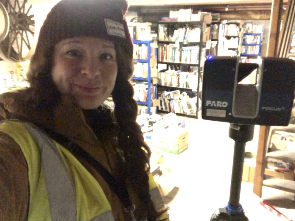 Archaeologist Kate Rogers stands next to a laser scanner on a tripod, with shelves and boxes of books behind her, and a large mill wheel to her right.