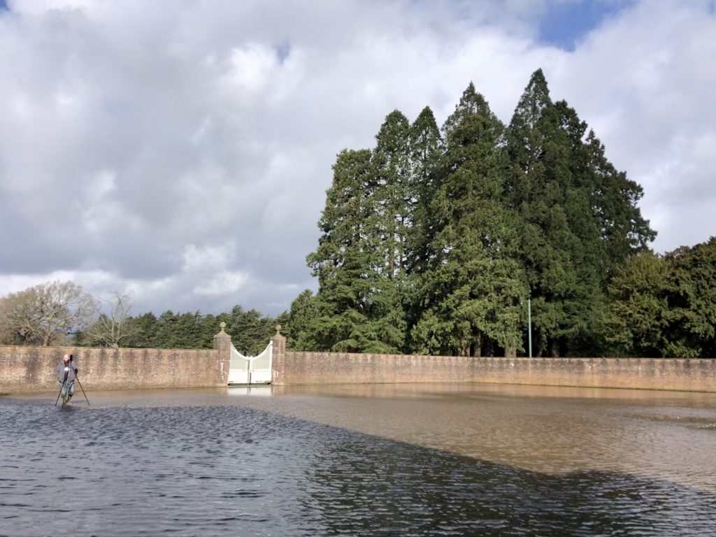 A view of a walled and gated outdoor courtyard the size of a football field. Archaeologist James Miles stands in the water at a distance with the laser scanner on a tripod.
