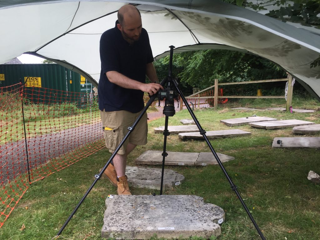 A man stands under a gazebo in the churchyard, operating camera on a tripod pointed directly down to photograph a headstone laid flat on the ground. There is a photographic key on the headstone and small black ball, and in his hand he holds a handheld camera flash.