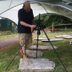 A man stands under a gazebo in the churchyard, operating camera on a tripod pointed directly down to photograph a headstone laid flat on the ground.