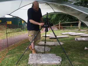 A man stands under a gazebo in the churchyard, operating camera on a tripod pointed directly down to photograph a headstone laid flat on the ground.