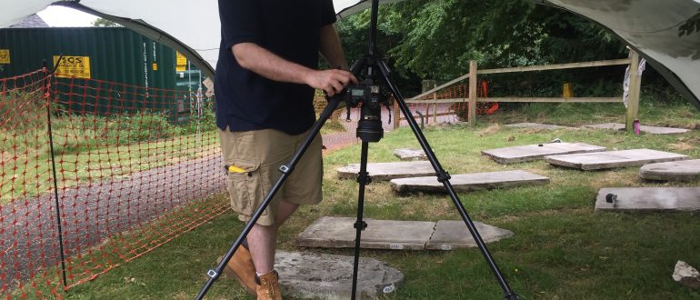A man stands under a gazebo in the churchyard, operating camera on a tripod pointed directly down to photograph a headstone laid flat on the ground.