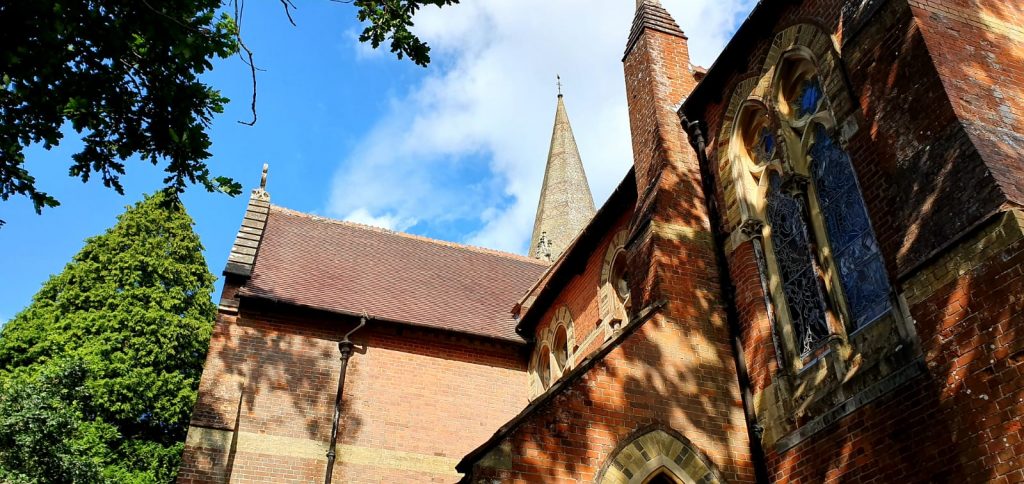 A view upwards of the windows and roof of a red brick church with a tall spire surrounded by trees.