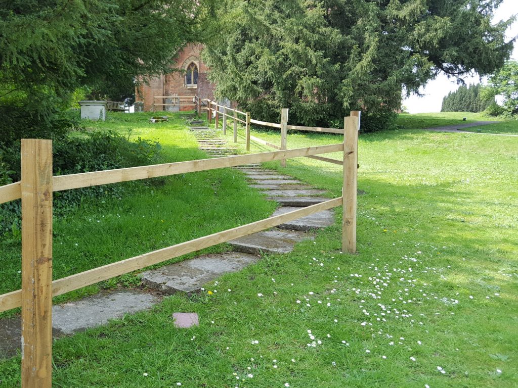 View of headstones laid flat as steps to make a path down a grassy green hillside from a red brick church. The path is fenced off from use by a wooden fence.