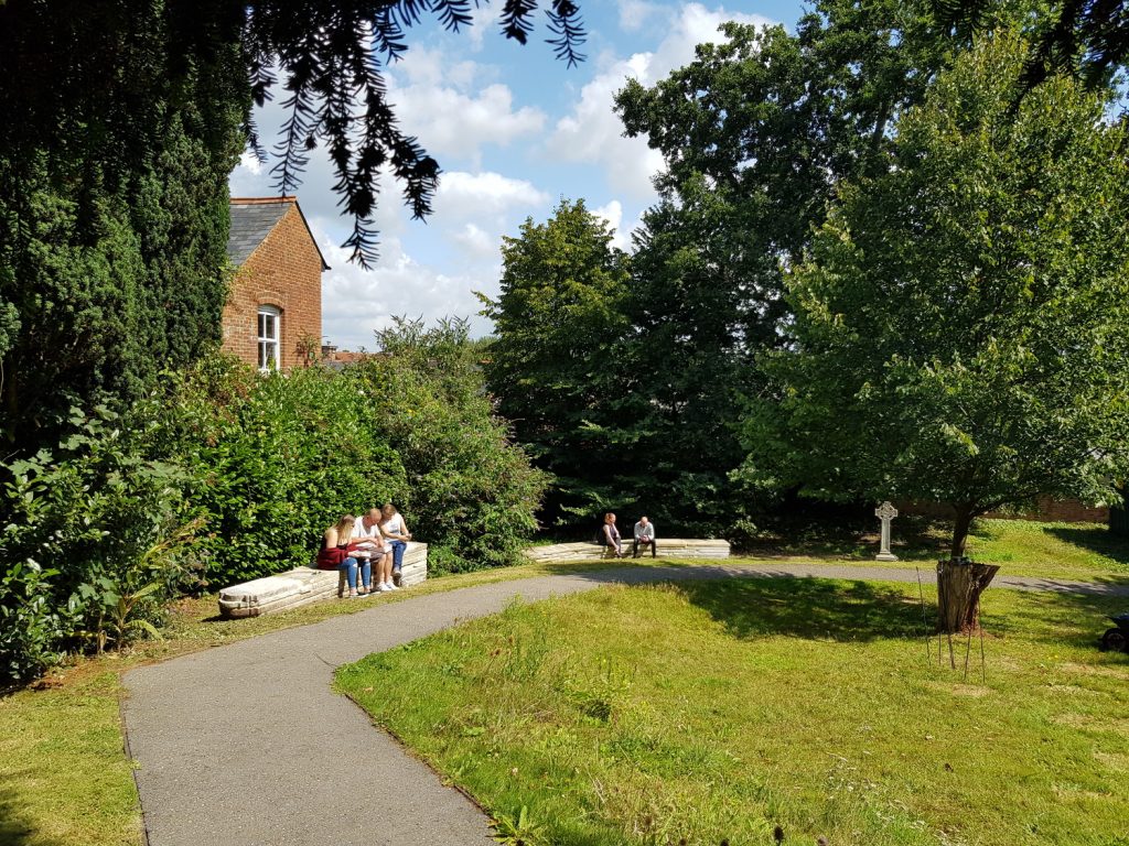 A view of the churchyrad after the project ended: the path is now made of tarmac and the headstones have been laid on top of and beside each other to form two long benches which people are sitting on.