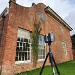 A laser scanner stands on a tripod outside an old redbrick building and manicured garden.