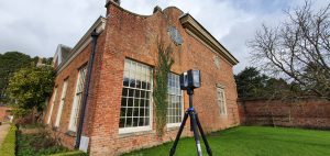 A laser scanner stands on a tripod outside an old redbrick building and manicured garden.