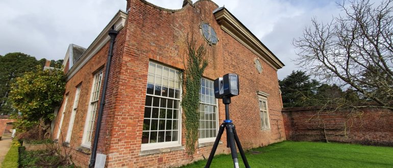 A laser scanner stands on a tripod outside an old redbrick building and manicured garden.